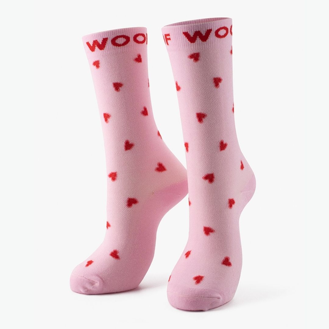 Studio shot of the pair of light pink Hearts Bamboo Crew Compression Socks standing upright, featuring all-over red mini heart motifs with bold red "WOOF" woven into the top cuffs.  